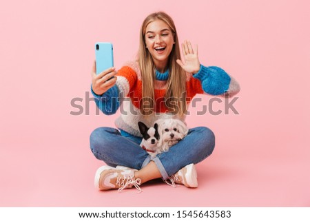Cute lovely girl wearing sweater sitting with legs crossed with her pet chihuahua and lapdog isolated over pink background