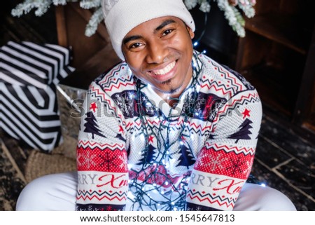Portrait of positive african-american man in xmas sweater and warm white hat holding LED lights on background of Christmas artificial tree with black balls and gift boxes. Dark color picture