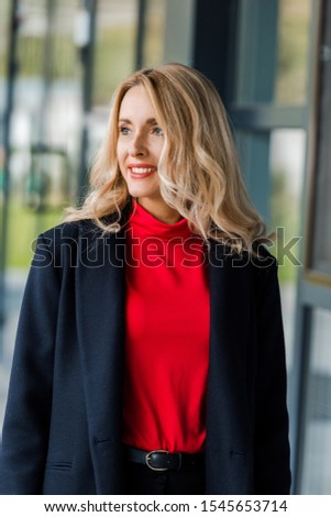 attractive and smiling businesswoman in black coat looking away 
