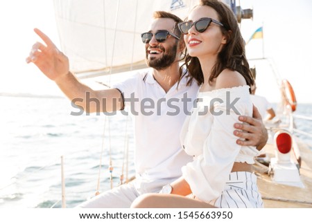 Image of a happy optimistic cheery young loving couple outdoors on yacht in sea pointing aside hugging.