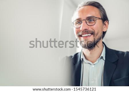 Photo of a Handsome focused and smiling young man sitting by the desk working at the computer. Blurry white computer screen visible on the left (might be used as copy space).