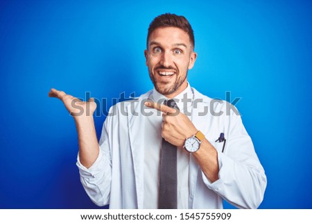 Young handsome doctor man wearing white professional coat over isolated background amazed and smiling to the camera while presenting with hand and pointing with finger.