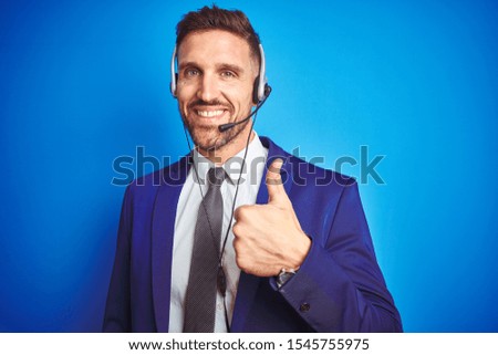 Young handsome operator man wearing call center headset over blue isolated background doing happy thumbs up gesture with hand. Approving expression looking at the camera with showing success.