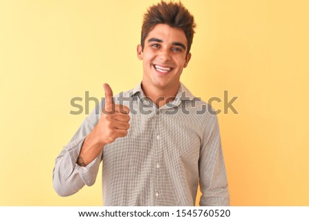 Young handsome man wearing casual shirt standing over isolated yellow background doing happy thumbs up gesture with hand. Approving expression looking at the camera with showing success.