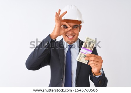 Young handsome architect man wearing helmet holding dollars over isolated white background with happy face smiling doing ok sign with hand on eye looking through fingers