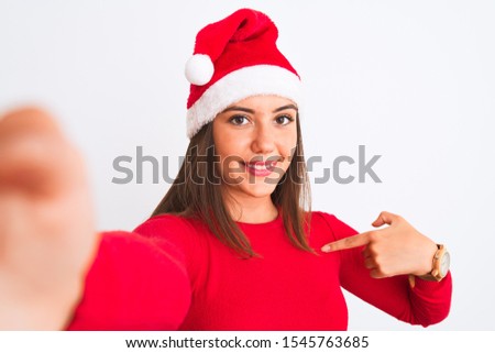 Girl wearing Christmas Santa hat make selfie by camera over isolated white background with surprise face pointing finger to himself