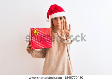 Beautiful redhead woman wearing christmas hat holding present over isolated background with open hand doing stop sign with serious and confident expression, defense gesture