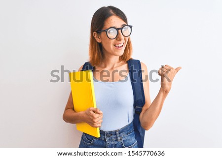 Beautiful redhead student woman wearing a backpack and book pointing and showing with thumb up to the side with happy face smiling