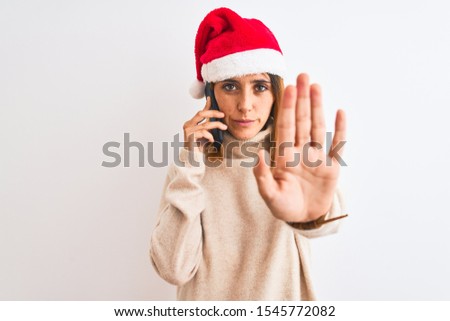 Beautiful redhead woman wearing christmas hat talking on smartphone with open hand doing stop sign with serious and confident expression, defense gesture