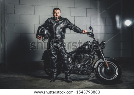Brutal bearded biker in leather suit is standing next to his bike while holding a helmet.