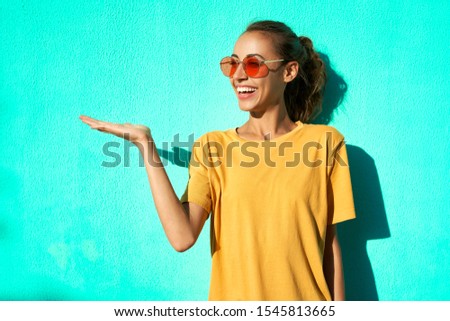 portrait of fashionable smilingl young woman in trendy red eyeglasses posing posing emotionally and smiling on blue background, wearing yellow t-short. Happy girl has attractive look