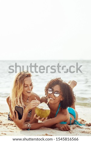 Two laughing young women wearing bikinis and drinking from a coconut taking selfies while suntanning on a sandy beach during vacation
