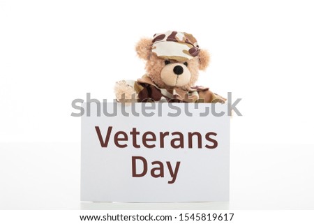 Veterans day concept. Cute teddy bear in soldier uniform holding a card with Veterans day text standing isolated against white background, copy space