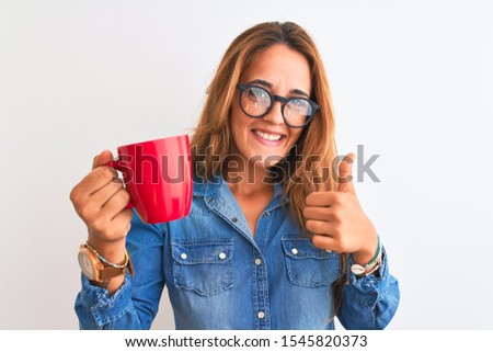 Young beautiful redhead woman wearing glasses drinking cup of coffee over isolated background happy with big smile doing ok sign, thumb up with fingers, excellent sign