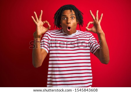Young afro man with dreadlocks wearing striped t-shirt standing over isolated red background looking surprised and shocked doing ok approval symbol with fingers. Crazy expression