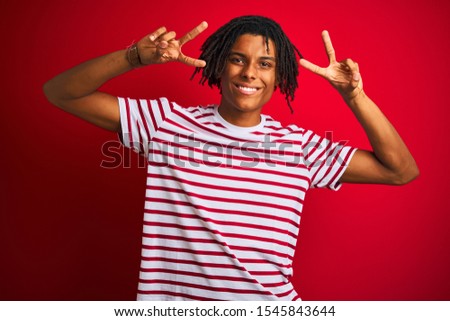Young afro man with dreadlocks wearing striped t-shirt standing over isolated red background Doing peace symbol with fingers over face, smiling cheerful showing victory