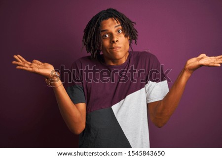Afro man with dreadlocks wearing casual t-shirt standing over isolated purple background clueless and confused expression with arms and hands raised. Doubt concept.