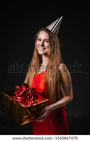 Pretty young woman in a festive cap holds a holiday box with a gift. Girl blonde is laughing and seductively looking at the camera. Shooting in professional studio on isolated black background.