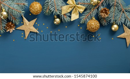 Christmas frame top border made of fir tree branches, golden decorative stars, balls over blue background. Flat lay, top view. Xmas banner mockup with copy space