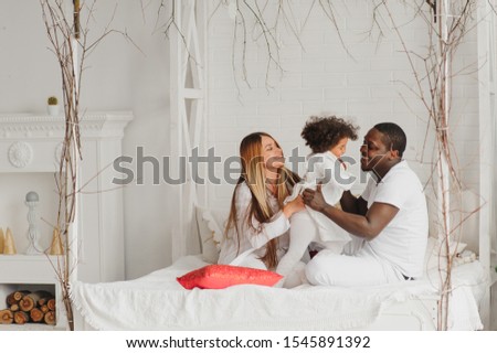 Portrait of happy multiracial young family lying on cozy white bed at home, smiling international mom and dad relaxing with little biracial girl child posing for picture in bedroom
