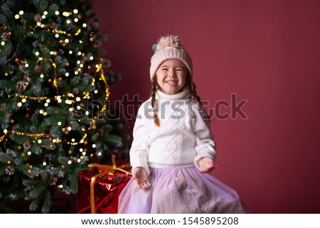 Beautiful little girl in the hat sitting near presents and christmas tree. Bokeh lights on the background. Concept christmas