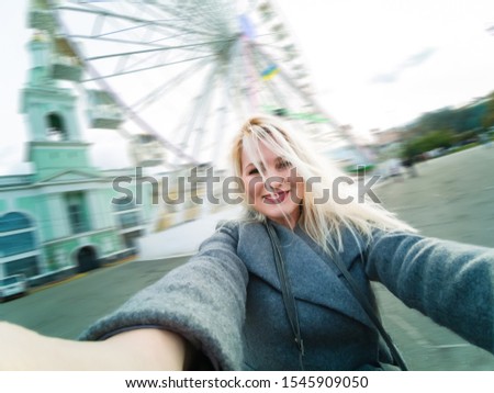 Attractive blonde girl in casual clothing, wearing a coat, takes selfie, looking into the camera. Stylish girl takes selfie, background of the ferris wheel