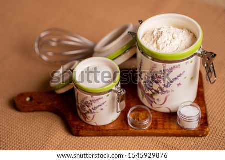 Top view of containers for flour and sugar on a chopping board and a hand whisk