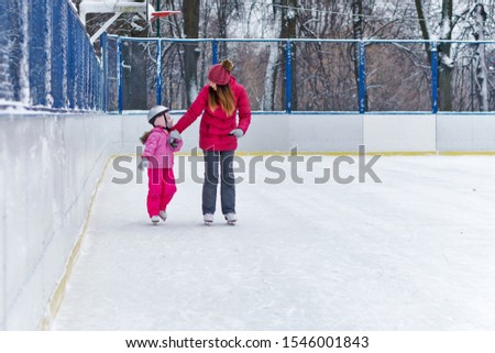 Mom teaches her little daughter to skate on the rink on a winter day. Weekends activities outdoor in cold weather.