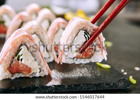 Macro shot of eating bacon uramaki sushi rolls with rice, cream cheese, smoked salmon, pork lard, cucumber, green onions, nori. Holding sushi rolls with chopsticks in Japanese restaurant closeup