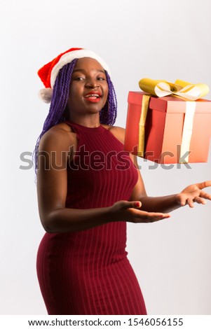 Beautiful Afro American girl in dress and Santa hat is holding presents, looking at camera and smiling, isolated on white background..