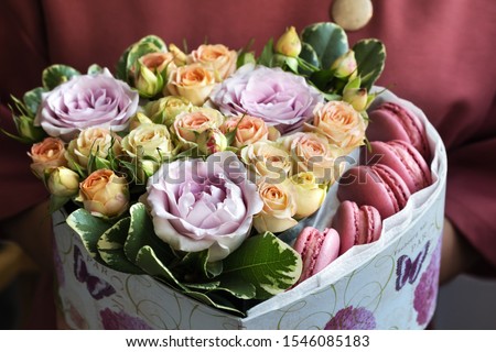 the girl is holding a box of flowers and sweets close-up with a blurred background. purple roses, pink roses, greens, pink macaroni. as a holiday gift, as a background