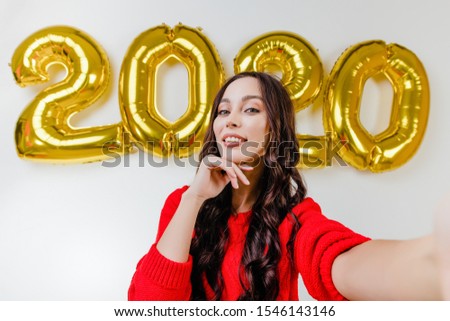 beautiful woman in red sweater making funny selfie in front of 2020 new year balloons