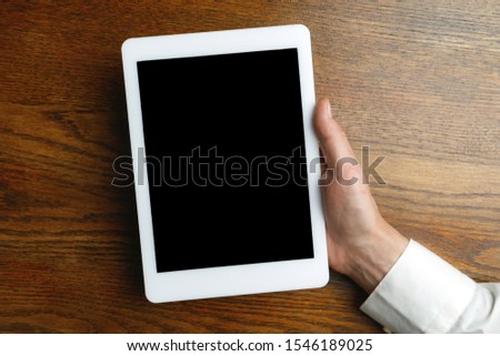 Male hand holding tablet with empty screen on wooden background for text or design. Blank gadget templates for contact or use in business. Finance, office, purchases. Mock up. Copyspace.