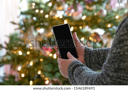  A woman is standing in front of decorated Christmas tree with her mobile phone. The display shows the Christmas tree and Copy Space. Only see the arms. Selective focus.                      