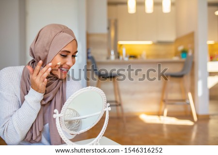Muslim woman spreading cream over her face while looking in the mirror. Beauty treatment. Female putting on moisturizer on her facial. Smiling Muslim woman applying cream to face and looking to mirror Royalty-Free Stock Photo #1546316252