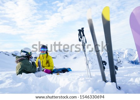 Picture of ski, snowboard, ski poles on the background of two tourists with thermos in hands sitting on snow resort.