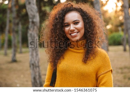 Photo of a positive cheery optimistic young curly woman walking in park outdoors.