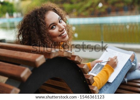 Photo of a positive beautiful young curly woman sit on bench in park outdoors writing notes in notebook.
