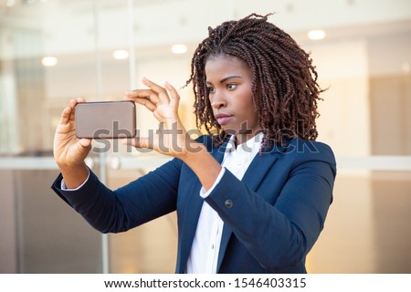 Focused female professional taking picture on cellphone outside. Young African American business woman standing near outdoor glass wall. Photo concept