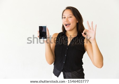 Woman showing smartphone and winking at camera. Beautiful cheerful young woman holding cell phone with blank screen and showing ok sign. Advertising concept