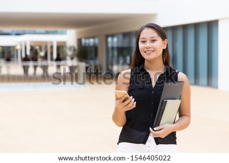 Cheerful female student holding notebooks and papers, texting message. Young Latin woman standing near city building, using mobile phone, looking at camera. Communication concept