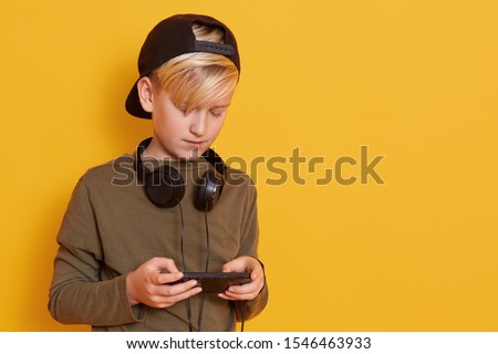 Picture of young boy with headphones around his neck, little guy wearing juniper green shirt and black back visor cap, holds his moder smartphone and playing online games. Copy space for advertisment.