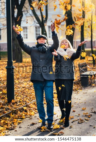 Happy blonde mature woman and handsome middle-aged brunette man walk in park and toss leaves. A loving couple of 45-50 years old walks in autumn park in warm clothes, vertical