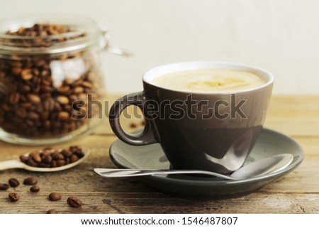 A cup of coffee and coffee seeds in a glass jar. Wooden table, light background. Horizontal orientation