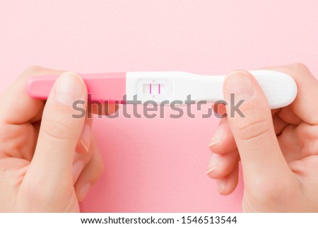 Young woman hands holding pregnancy test with two stripes on pastel pink background. Positive result. Closeup. Point of view shot. 