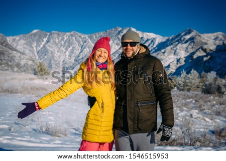 Young beautiful couple in bright clothes on the background of snowy mountains on winter sunny day. Guy and girl during new year holidays in the mountains. Cheerful people smile and look at camera.