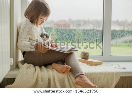 cute little girl sitting near the window, reading a book and holding a gray kitten in her arms Royalty-Free Stock Photo #1546521950