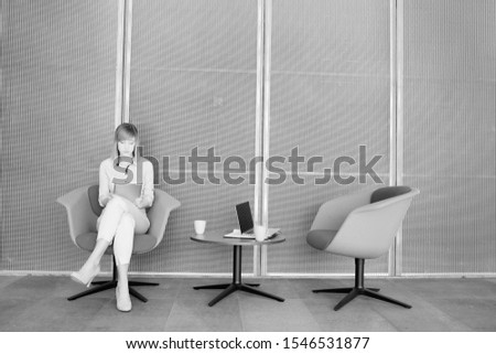 Black and white photo of businesswoman using digital tablet while waiting and drinking coffee or tea in office lobby