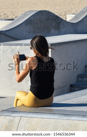 back of attractive caucasian woman taking pictures with her cell phone brunette hair black tank top yellow tight pants sitting on cement with decorative cement background