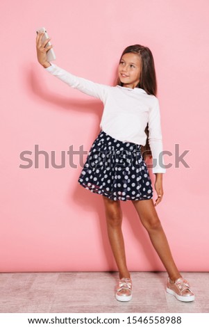 Photo of optimistic young girl child posing isolated over pink wall background take a selfie by mobile phone.